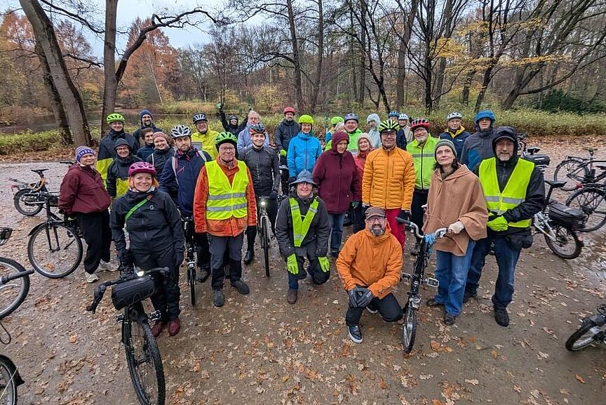ADFC-Gruppe bei einer herbstlichen Radtour Gruppenfoto von gut gelaunten Radfahrenden in wetterfester Kleidung, die mit ihren Fahrrädern auf einem laubbedeckten Waldweg stehen. Viele tragen Fahrradhelme und einige gelbe Warnwesten. Im Hintergrund sind herbstliche Bäume und ein bewölkter Himmel zu sehen.